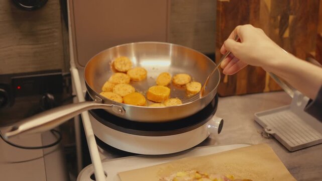 Close-up of woman hands using fork to flip sizzling sweet potato slices in hot frying pan on induction stove. Food prep and cooking process on stainless steel countertop in warm home kitchen.