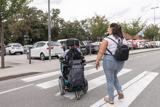 A man in a wheelchair is assisted by a woman while walking on a city street. They are near parked cars and trees.