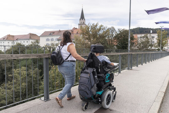 A woman helps a man in a wheelchair as they navigate a bridge in an urban area. They enjoy their time outdoors, fostering independence.