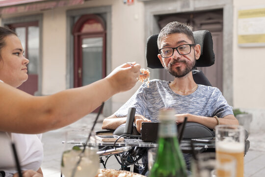 A man in a wheelchair enjoys a meal with his personal assistant at a restaurant. They are focused on each other while dining.