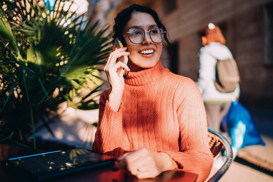 Happy hispanic woman phoning with smartphone and smiling, joyful mobile conversation in city cafe, looking away, authentic representation, positive attitude, connection and digital lifestyle.
