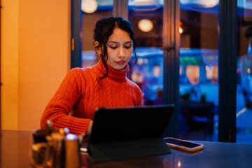 Hispanic woman focused on working with digital tablet in modern cafe setting, young female professional typing on mobile device at night, authentic digital nomad lifestyle and urban connectivity. © BullRun