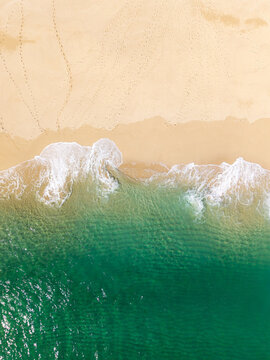 Aerial view directly above soft white waves lapping a golden sandy beach