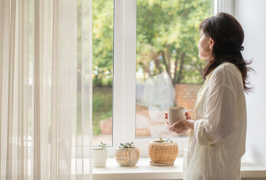 Modern adult woman in simple loungewear stands by window with cup of coffee, enjoying quiet moment of solitude. A slow morning ritual, an authentic moment in life of real person