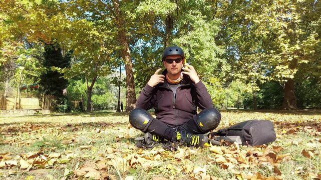 Man putting on and securing a protective helmet strap while sitting on park grass with roller skates on his feet, preparing for a safe workout. Commercial B-roll, 4K UHD 