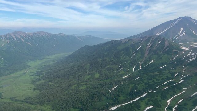 Aerial view from helicopter over dramatic mountain ridges, green valleys and lingering snowfields in Kamchatka. Scenic flight, pristine wilderness, volcanic landscape, adventure travel, nature tourism