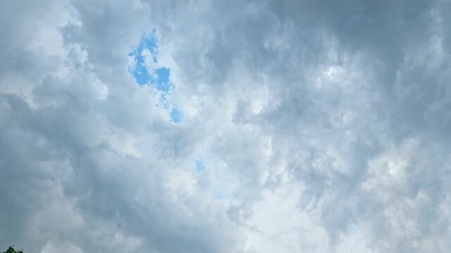 This panning video clip captures a dynamic overcast sky filled with heavy storm clouds, interspersed with patches of bright blue sky, trees, a street lamp, and solar panels. 