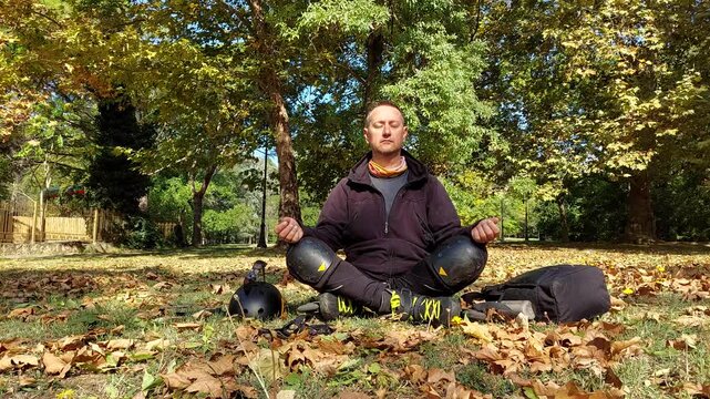 Man wearing inline skates and protective knee pads sitting cross-legged on green lawn with fallen leaves in a sunny autumn park, meditating and breathing deeply during a workout break. Commercial 4k