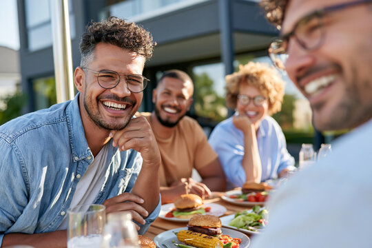 Sunlit table, smiling friends enjoying burgers and drinks. Laughing, leaning in, capturing warm, casual connection. Modern architecture frames the cheerful, relaxed gathering. Perfect for lifestyle