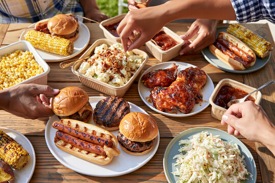 Hands reach for food at a lively outdoor gathering. Burgers, sausages, corn, and coleslaw fill the table. Friends enjoy a warm, casual meal together. Sunlight bathes the table