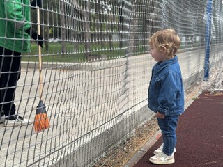 Toddler girl in denim jacket looking thoughtful near sports ground fence © Nastya