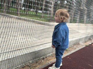 Toddler girl in denim jacket looking thoughtful near sports ground fence © Nastya