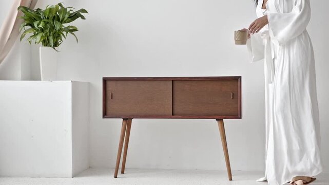 Elegant Woman in White Robe Placing a Ceramic Mug on a Mid-Century Modern Wooden Sideboard in a Minimalist Bright Interior