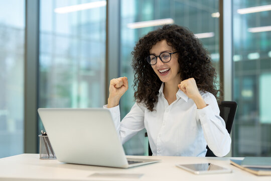 Young professional businesswoman in glasses showing excitement and celebrating a triumph or achievement with clenched fists while working on a laptop in a modern office