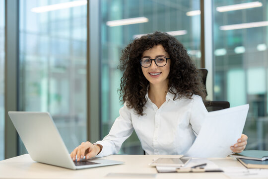 Young professional businesswoman smiling at the camera while working with a laptop and holding paperwork at a modern office desk, embodying efficiency and corporate success