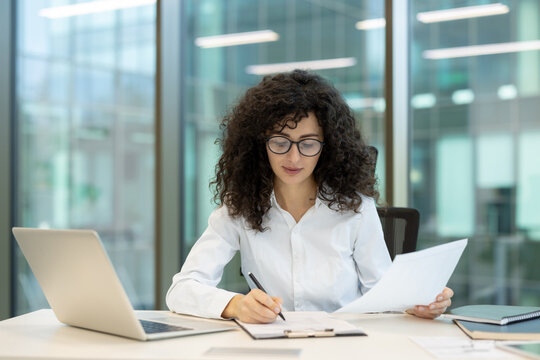 Businesswoman with curly hair and glasses sitting at a white office desk, focusing on writing and reviewing paperwork while a laptop sits nearby, conveying concepts of productivity and organization