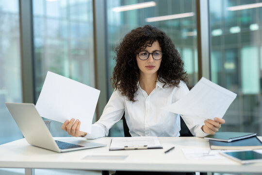 Woman with curly hair and glasses sitting at an office desk, holding multiple documents and looking stressed while dealing with work overload and complicated paperwork