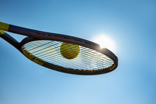 Close up of a tennis ball resting on racket strings against a bright sky