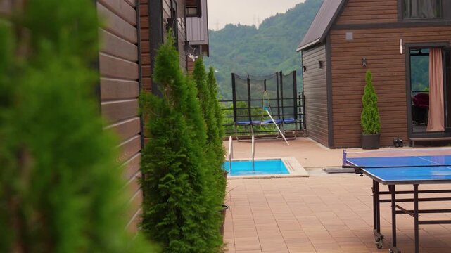 Beautiful view of a modern wooden cottage house with a private swimming pool and a patio featuring a ping-pong table and trampoline, all set against a stunning mountain forest backdrop