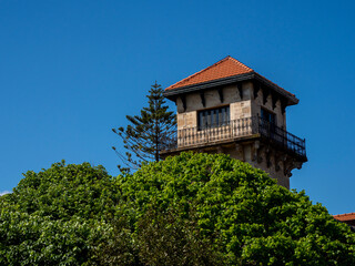 Historic Stone Tower With Red Tile Roof - Elegant historic stone tower with ornate iron balcony and terracotta roof rising above lush green treetops against a clear blue sky. © jmag.foto