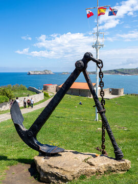 Large Anchor Monument on Spanish Coast - Historic black anchor monument on a grassy hill overlooking the turquoise sea with islands and flags under a bright blue sky in Spain.