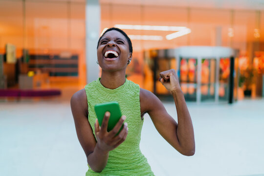 Laughing dark skinned model rejoicing successful online goal completion via mobile app. Overjoyed African American woman raising fist, experiencing pure happiness with smartphone in terminal.