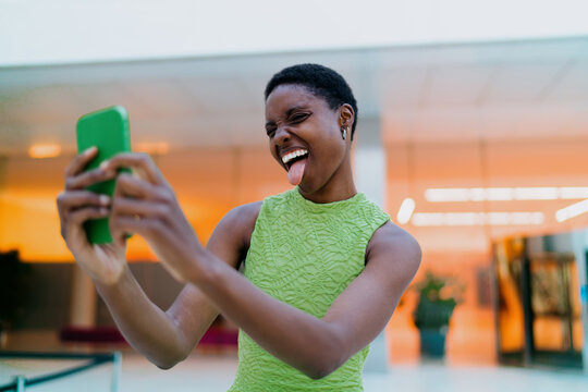 Joyful African American woman making selfie video call on phone screen. Happy black female influencer waving hand or recording funny vlog via smartphone camera software application indoors.