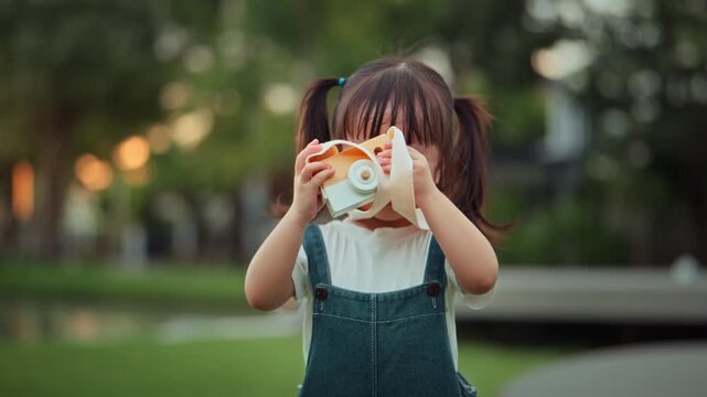 Cute Asian toddler girl using wooden toy camera outdoors in green park, little child pretending to take photos while learning photography, creativity, curiosity, imagination and childhood exploration 