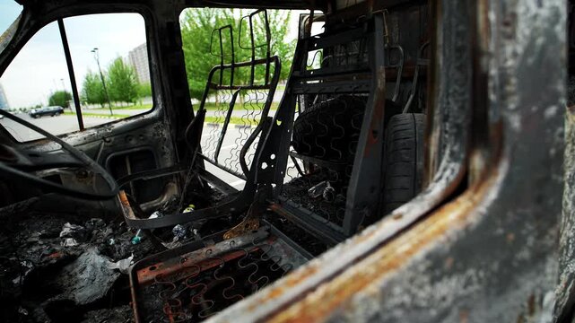 Charred automobile cabin shows severe destruction across driver seat and dashboard area. Burned vehicle demonstrates aftermath of intense fire leaving rusted metal frame exposed to elements.