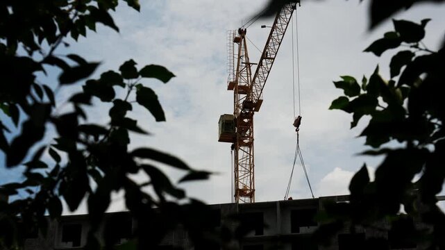 Industrial tower crane works steadily at urban construction development zone. Tall machinery towers over concrete structure while dark tree foliage creates dynamic natural foreground framing outdoors.