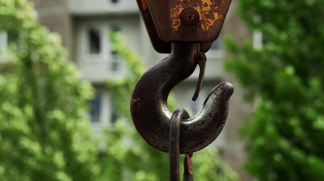 Industrial metal crane hook swings slightly against urban architecture backdrop. Sturdy lifting equipment remains suspended outdoors before tall apartment complex surrounded by summer greenery.