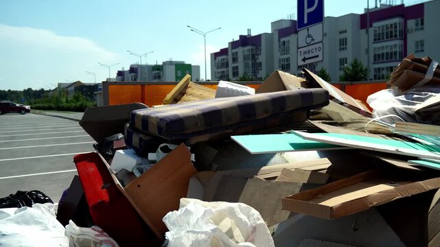 Discarded household furniture and cardboard clutter large open top dumpster outdoors. Piles of bulky waste lie disorganized inside waste bin near modern apartment buildings under bright sky.