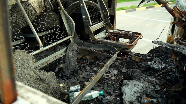 Burned passenger van cabin displays charred debris and melted fixtures following severe fire. Ruined vehicle stands abandoned outdoors with charred framework and ash covering floorboards.