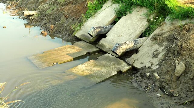 Drainage ditch receives water runoff from twin metal pipes resting on concrete blocks. Murky water flows steadily through outdoor canal channel flanked by green grass and earthen banks.