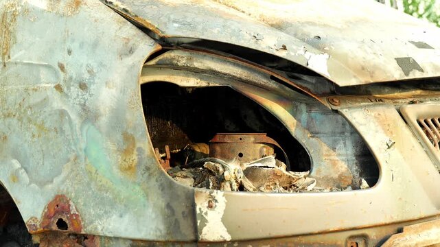 Charred automobile body shows extensive corrosion, featuring broken headlight compartment and burnt structural frame. Rusted motor vehicle remains stationary under bright natural sunlight.