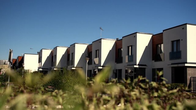 Row of modern suburban townhouses stands under clear blue sky behind soft green foliage in foreground. Contemporary residential neighborhood creates peaceful and inviting atmosphere outside.