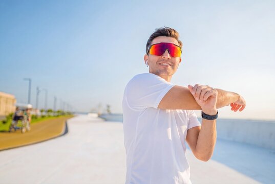 Athletic man stretching arms on sunny boardwalk, fit male runner warming up outdoors, healthy lifestyle training in summer sun.