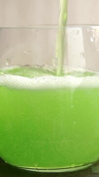 Macro close-up video showing fresh green herbal or lime juice pouring into a clean transparent glass and forming sparkling fizzy bubbles under bright morning sunlight, ideal for wellness and detox bev