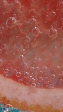 Close-up vertical macro video showing a fresh pink grapefruit slice submerged in sparkling carbonated water covered in tiny bubbles.