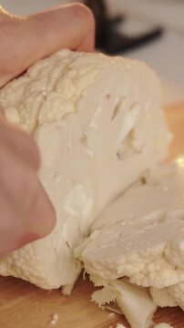 Cook slices whole cauliflower head on wooden cutting board with knife for raw food diet cuisine. Natural lighting highlights fresh organic vegetable during healthy kitchen meal preparation.