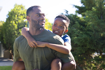 Fototapeta premium Father carrying youth son piggyback in yard wearing olive-green and blue tees smiling toward right