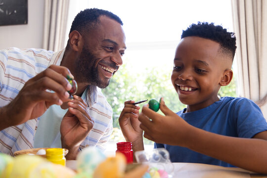 African American father and child painting eggs with paint jars and brushes at table by window