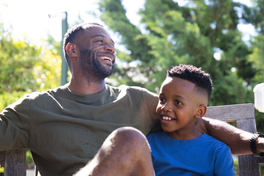 African American father and son laughing, sitting on bench in sunny park wearing t-shirts and watch