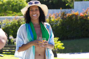 African American woman standing poolside in green swimwear straw hat holding orange cocktail glass