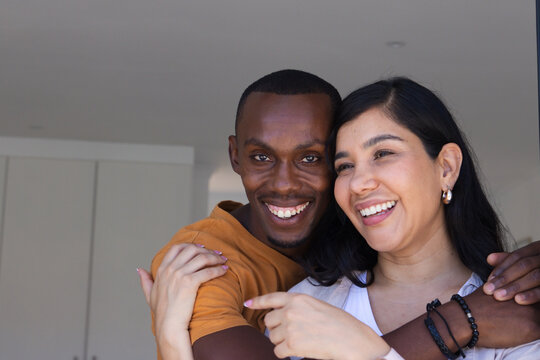 Diverse couple embracing and smiling while woman wearing lavalier mic and bracelets at home