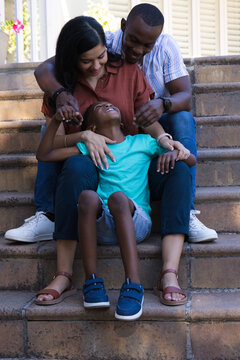 Family sitting on front stone steps, woman holding laughing child, potted pink flowers nearby