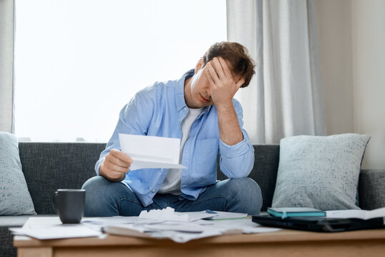 stressed man sits holding unpaid utility bills with severe financial stress from high inflation, rising energy costs, and heavy household expense debts during an economic crisis.