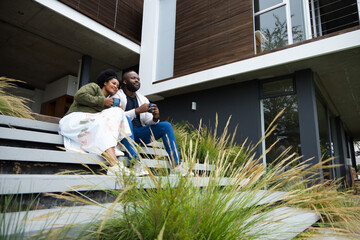 Obraz premium Couple sitting on wide horizontal steps outside modern two-story house, holding ceramic mugs