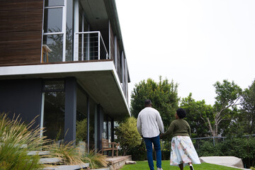 African American couple walking on lawn toward modern glass home, passing stone steps, copy space