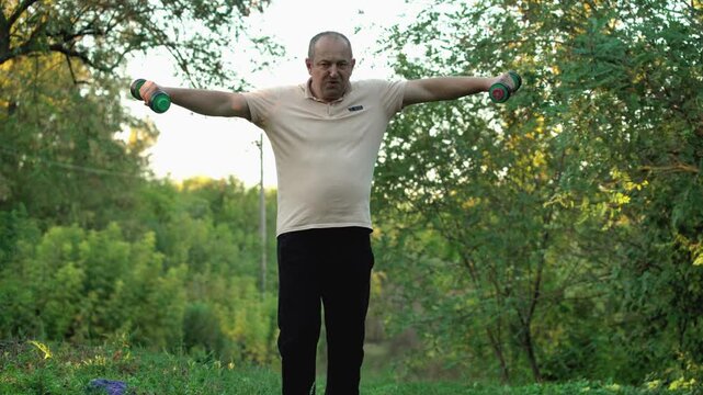 Older adult performing dumbbell lateral raises while standing on a forest path surrounded by green trees and natural foliage during outdoor exercise in daylight